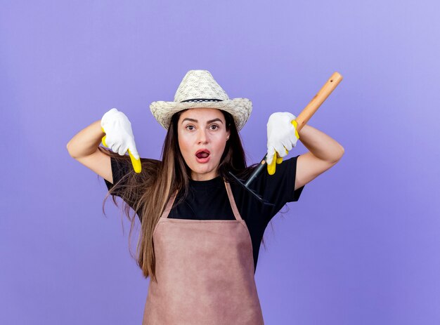 Surprised beautiful gardener girl in uniform wearing gardening hat and gloves holding rake and points at down isolated on blue background