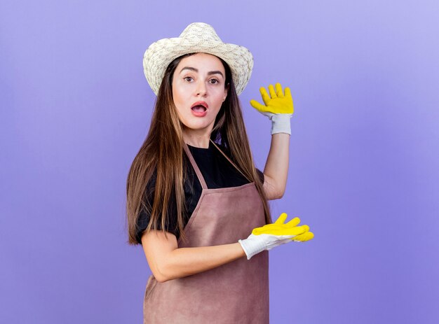 Удивленная красивая девушка-садовник в униформе в садовой шляпе и перчатках указывает руками на бок, изолированными на синем фоне