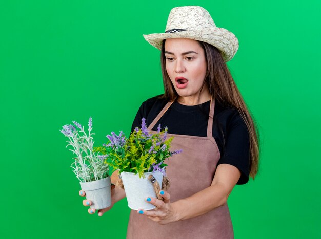 Surprised beautiful gardener girl in uniform wearing gardening hat holding and looking at flower in flowerpot isolated on green background