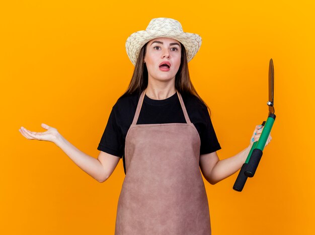 Surprised beautiful gardener girl wearing uniform and gardening hat holding clippers and spread hand isolated on orange background