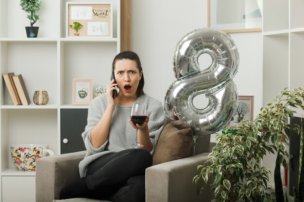 Surprised beautiful girl on happy women day holding glass of wine speaks on wine sitting on armchair in living room