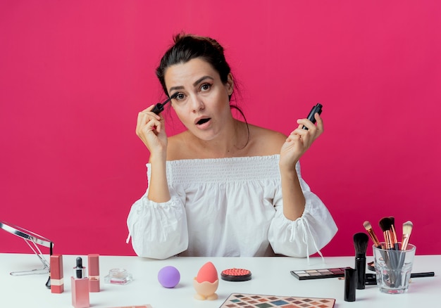 Free photo surprised beautiful girl sits at table with makeup tools applying mascara looking isolated on pink wall