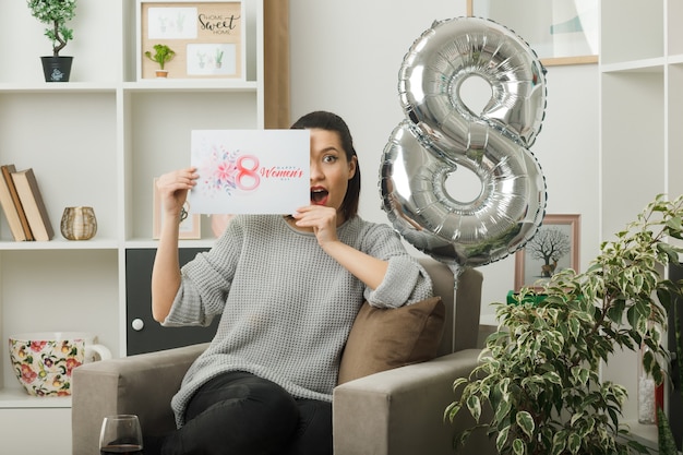 Bella donna sorpresa durante la giornata delle donne felici che tiene in mano e copre il viso con una cartolina seduta su una poltrona in soggiorno