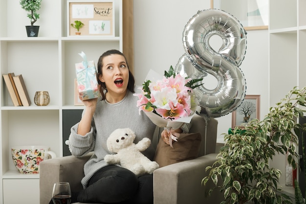 Free photo surprised beautiful woman on happy women day holding present with bouquet sitting on armchair in living room