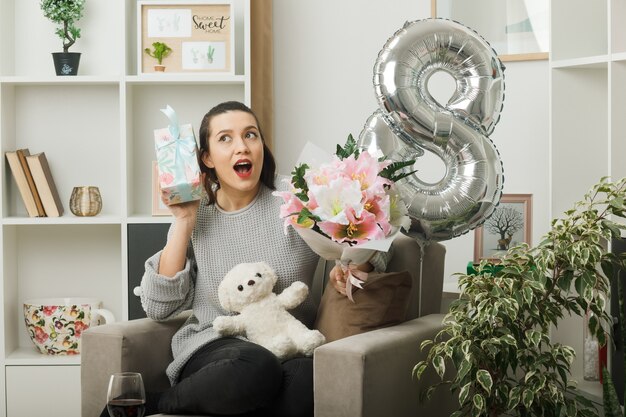 Surprised beautiful woman on happy women day holding present with bouquet sitting on armchair in living room