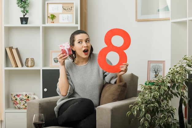 Surprised beautiful woman on happy women day holding present with number eight sitting on armchair in living room