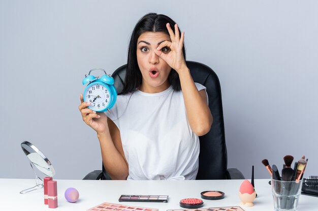 Surprised beautiful woman sits at table with makeup tools holding alarm clock showing look gesture 