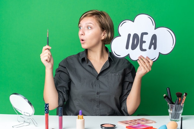 Free photo surprised beautiful woman sits at table with makeup tools holding idea bubble with makeup brush