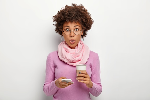 Free photo surprised beautiful woman with crisp hair checks newsfeed, gets shocked message, holds takeout coffee, cannot believe in something, wears optical glasses and purple sweater, poses indoor