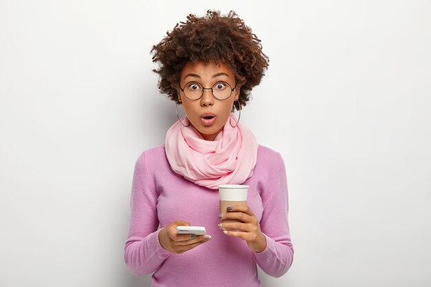 Surprised beautiful woman with crisp hair checks newsfeed, gets shocked message, holds takeout coffee, cannot believe in something, wears optical glasses and purple sweater, poses indoor