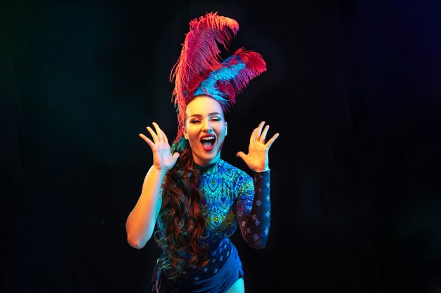 Surprised. Beautiful young woman in carnival, stylish masquerade costume with feathers on black background in neon light.