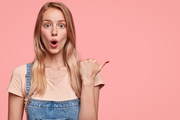 Surprised beautiful young woman with shocked expression, points aside with thumb, keeps jaw dropped, dressed in denim overalls