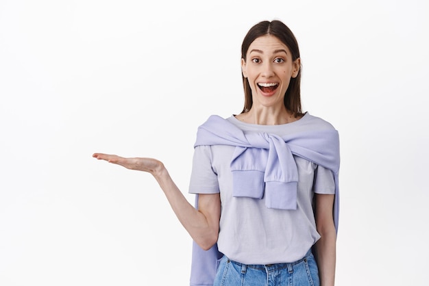 Surprised beauty girl holding empty space in open hand display at item on her palm look amazed at camera standing over white background