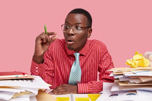 Surprised black businessman makes tiny gesture, being astonished with low profit, dressed in pink striped shirt, tie, works with documentation