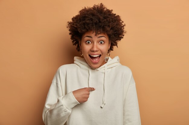 Surprised black woman points at herself, shocked be chosen or picked as leader of team, wears white sweatshirt, keeps mouth opened