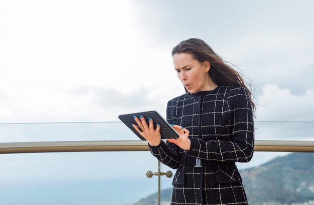 Surprised blogger girl is looking at planshet computer in hand against the background of city view