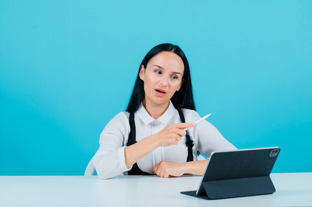 Surprised blogger girl is pointing right with pencil by sitting in front of tablet camera on blue background