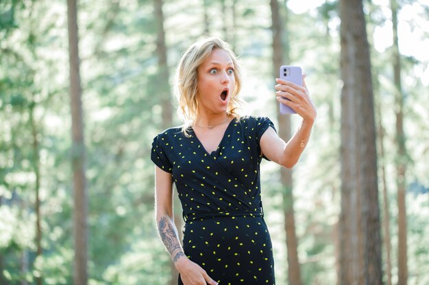 驚いたブロガーの女の子は、自然の背景に彼女の携帯電話でセフィーを取っています