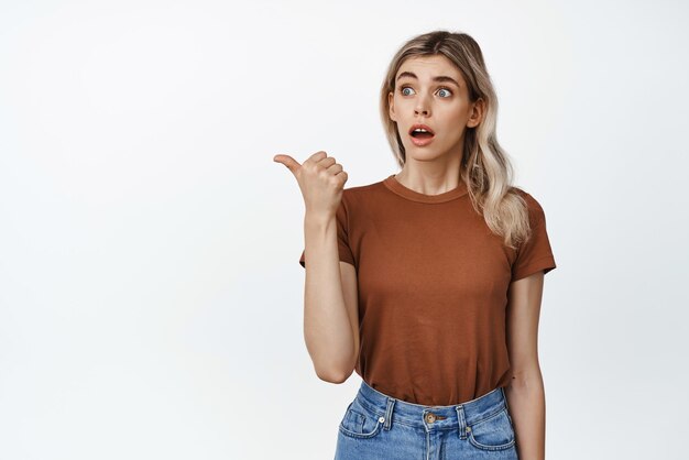 Surprised blond girl gasp impressed pointing finger left and looking at advertisement amazed standing against white background