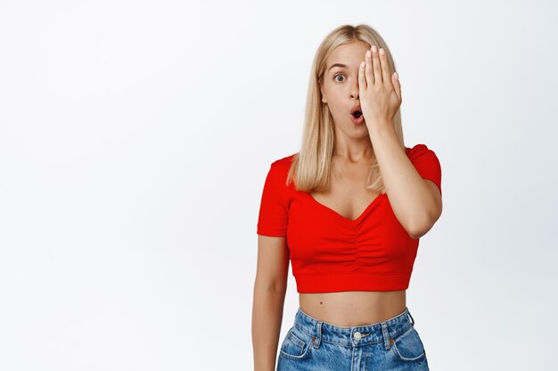 Surprised blond woman gasping covering half face looking with one eye with shocked face expression standing against white background