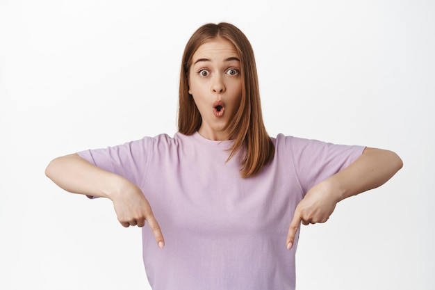 Surprised blond woman, woman showing advertisement, pointing down, say wow and gasp in awe, standing impressed against white wall.