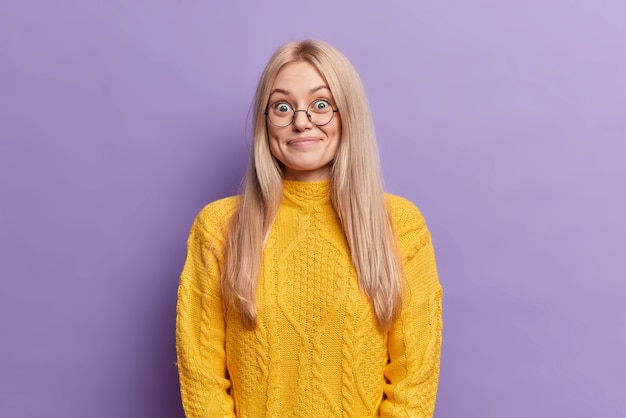 Surprised blonde European woman has cheerful expression wears round spectacles hears unexpected pleasant news dressed in yellow sweater 