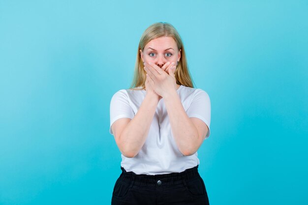 Surprised blonde girl is holding hands on mouth on blue background