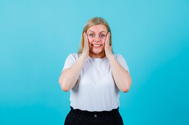 Surprised blonde girl is looking at camera by putting hands on cheeks on blue background