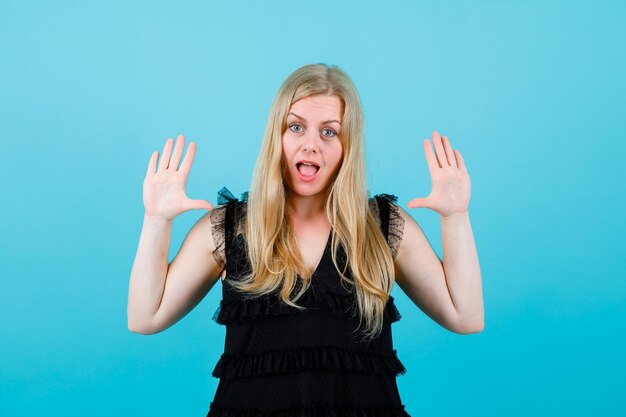 Surprised blonde girl is looking at camera by raising up her handfuls on blue background