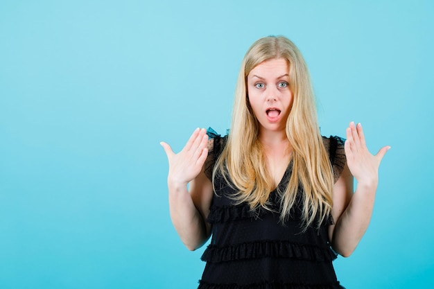 Free photo surprised blonde girl is looking at camera by raising up her hands on blue background