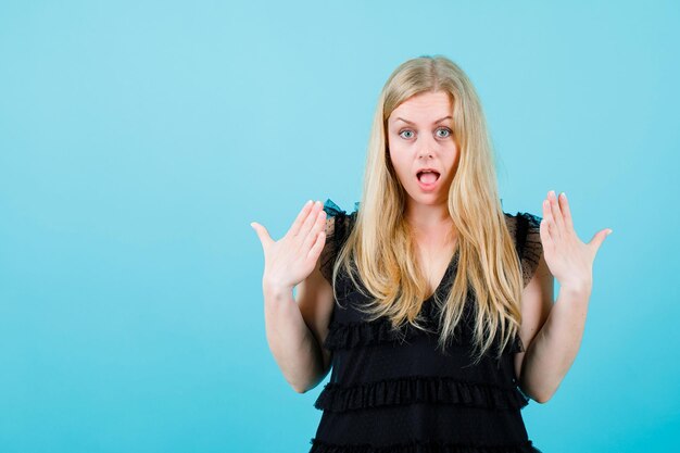 Surprised blonde girl is looking at camera by raising up her hands on blue background