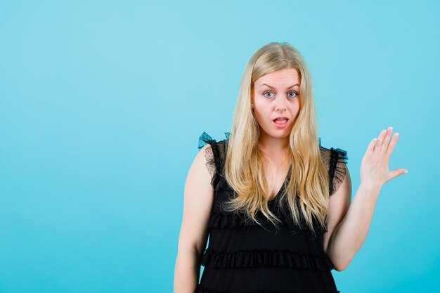 Surprised blonde girl is raising up and showing her hand on blue background