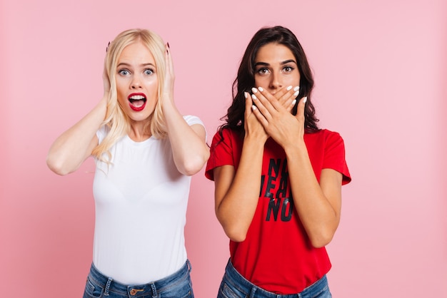 Surprised Blonde woman covering her ears and brunette woman covering her mouth over pink 