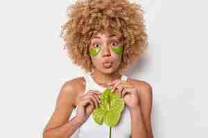 Foto gratuita la donna bionda sorpresa mantiene le labbra arrotondate tiene il fiore di calla applica cerotti di idrogel verde sotto gli occhi per rimuovere il gonfiore vestito con una maglietta casual isolato su sfondo bianco concetto di benessere