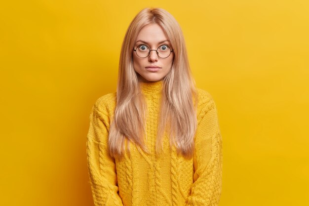 удивленная блондинка стоит безмолвно в помещении, носит круглые прозрачные очки, одетый в желтый свитер, позирует в помещении. Удивленная впечатленная европейская женщина смотрит искривленными глазами.