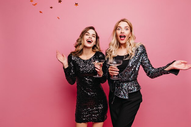 Surprised blonde woman with red lipstick standing under confetti  Graceful trendy ladies posing on rosy wall during party.