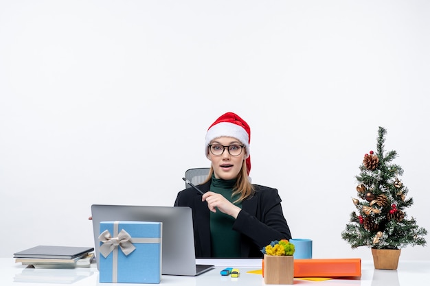 Surprised blonde woman with a santa claus hat sitting at a table with a Christmas tree and a gift on it and pointing something on the right side on white background