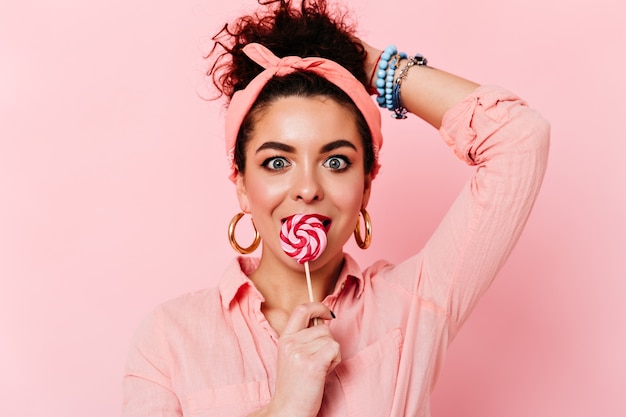 Surprised blue-eyed brunette girl in pink pin-up outfit eats lollipop on isolated space.