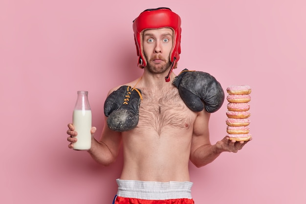 удивленный голубоглазый мужчина смотрит в защитной шляпе, боксерские перчатки на шее, голый торс, держит бутылку с молоком, а куча пончиков испытывает искушение съесть нездоровую пищу