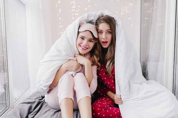 Surprised brown-haired woman in red pyjamas sitting in cozy bedroom. Indoor shot of refined caucasian girl hiding under white blanket.