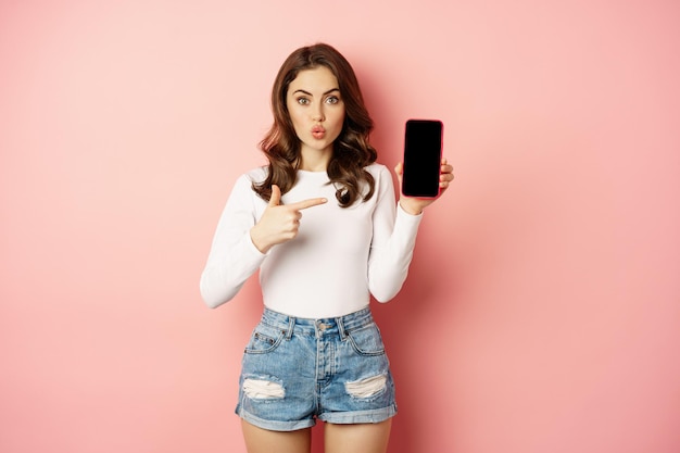 놀란 브루네트 소녀는 휴대폰 앱 화면을 가리키며 스마트폰 광고, 온라인 쇼핑 제안을 보여주고 분홍색 배경에 놀란 채 서 있습니다.