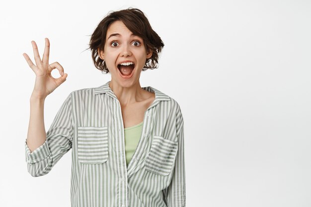 Surprised brunette girl shows okay, ok sign, look amazed, praise smth good, positive feedback, standing on white.