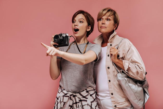 Surprised brunette lady in t-shirt points finger to side, holds camera and poses with old woman with bag in light clothes on pink backdrop.