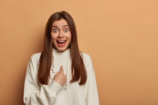 Surprised brunette millennial girl happy being chosen, points at herself, gazes at camera, wears white sweatshirt, smiles positively