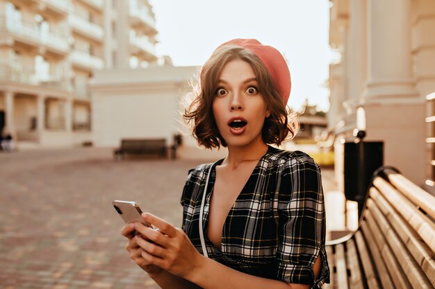 Surprised brunette woman in elegant blouse sitting on bench. Shocked white girl in trendy beret holding her smartphone.