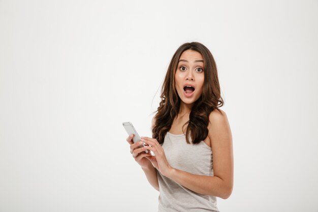 Surprised brunette woman holding smartphone and looking at the camera with open mouth over gray