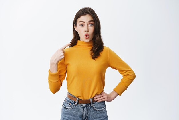 Surprised brunette woman say wow pointing at herself and staring at camera with disbelief being chosen or named standing on white background