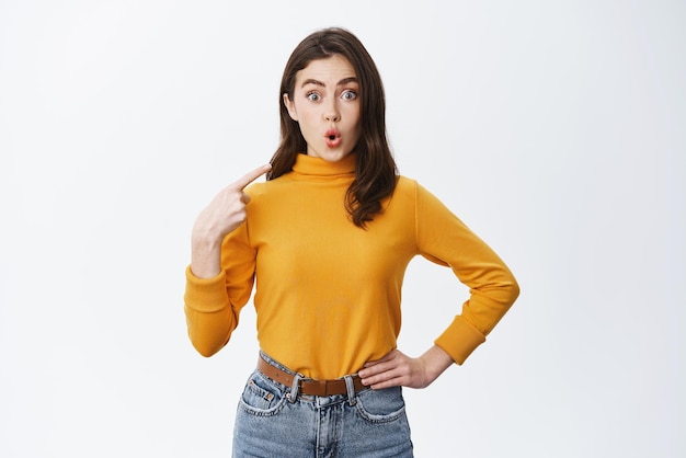 Surprised brunette woman say wow pointing at herself and staring at camera with disbelief being chosen or named standing on white background