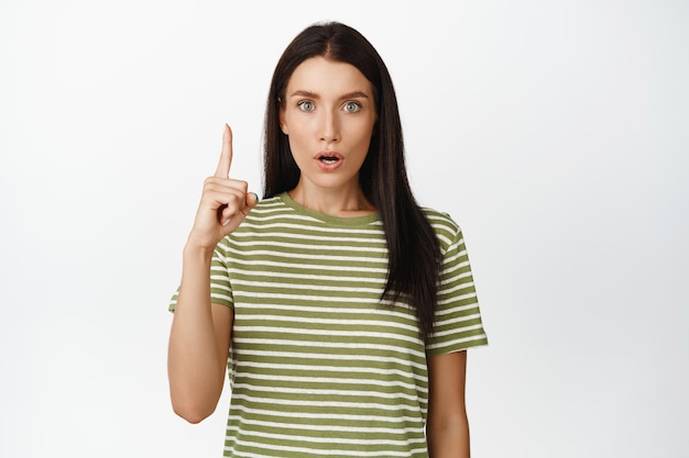 Surprised brunette woman staring impressed at camera pointing finger up amazed by something upwards standing over white background