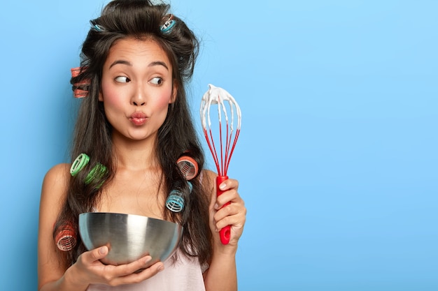 Surprised brunette woman whisks sour cream in bowl, wears hair curlers